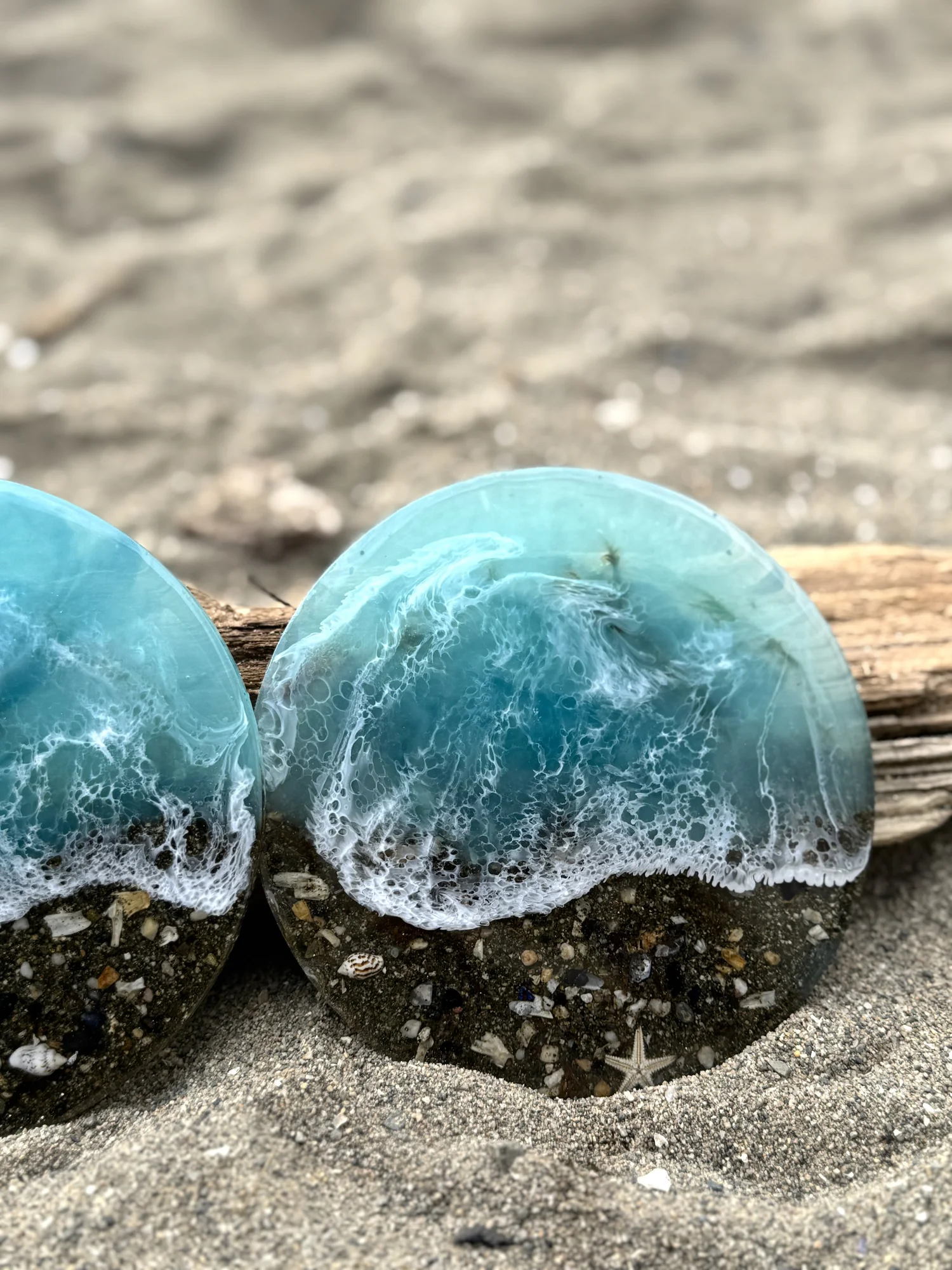 Two decorative glass sea wave sculptures placed on the sand near a piece of driftwood.