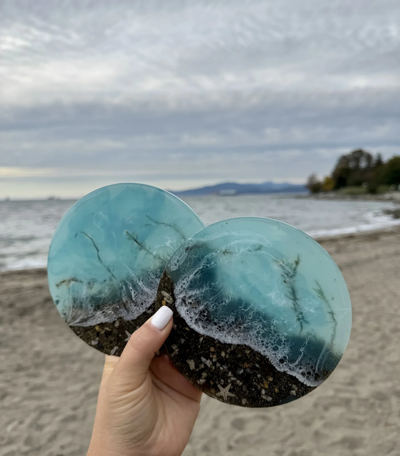 Two resin coasters designed to look like ocean waves with shells and sand inside, held up by a hand on a beach with water and cloudy sky in the background.