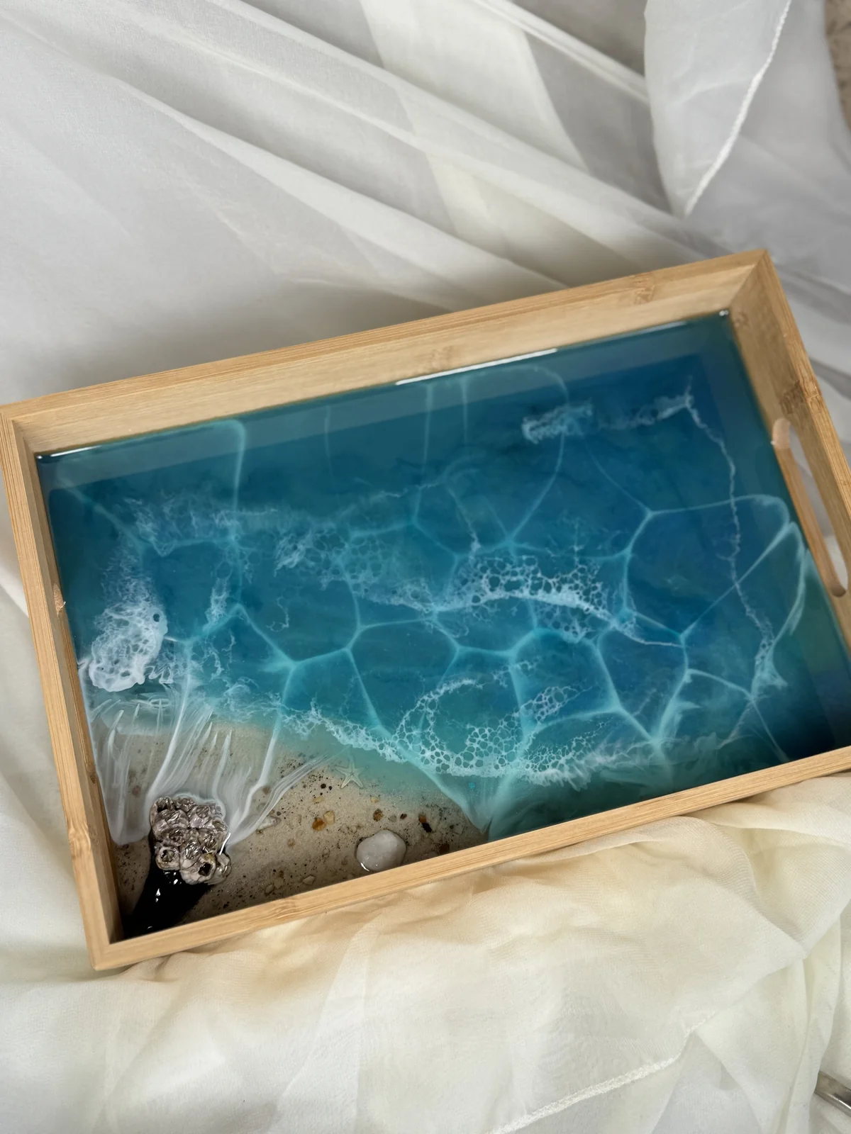 A wooden tray with a resin art piece designed to look like ocean waves crashing onto a beach, featuring a sandy area with small rocks and a metallic seashell.