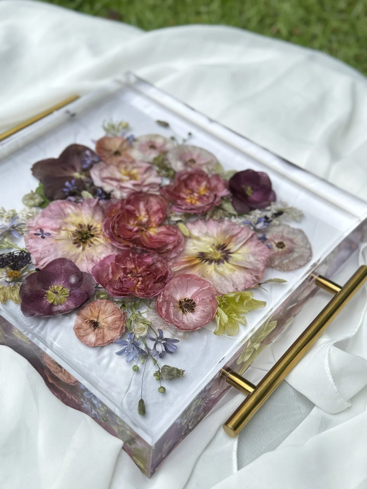 A transparent box filled with pressed pink, purple, and yellow flowers, placed on white fabric with a gold handle visible in the foreground.