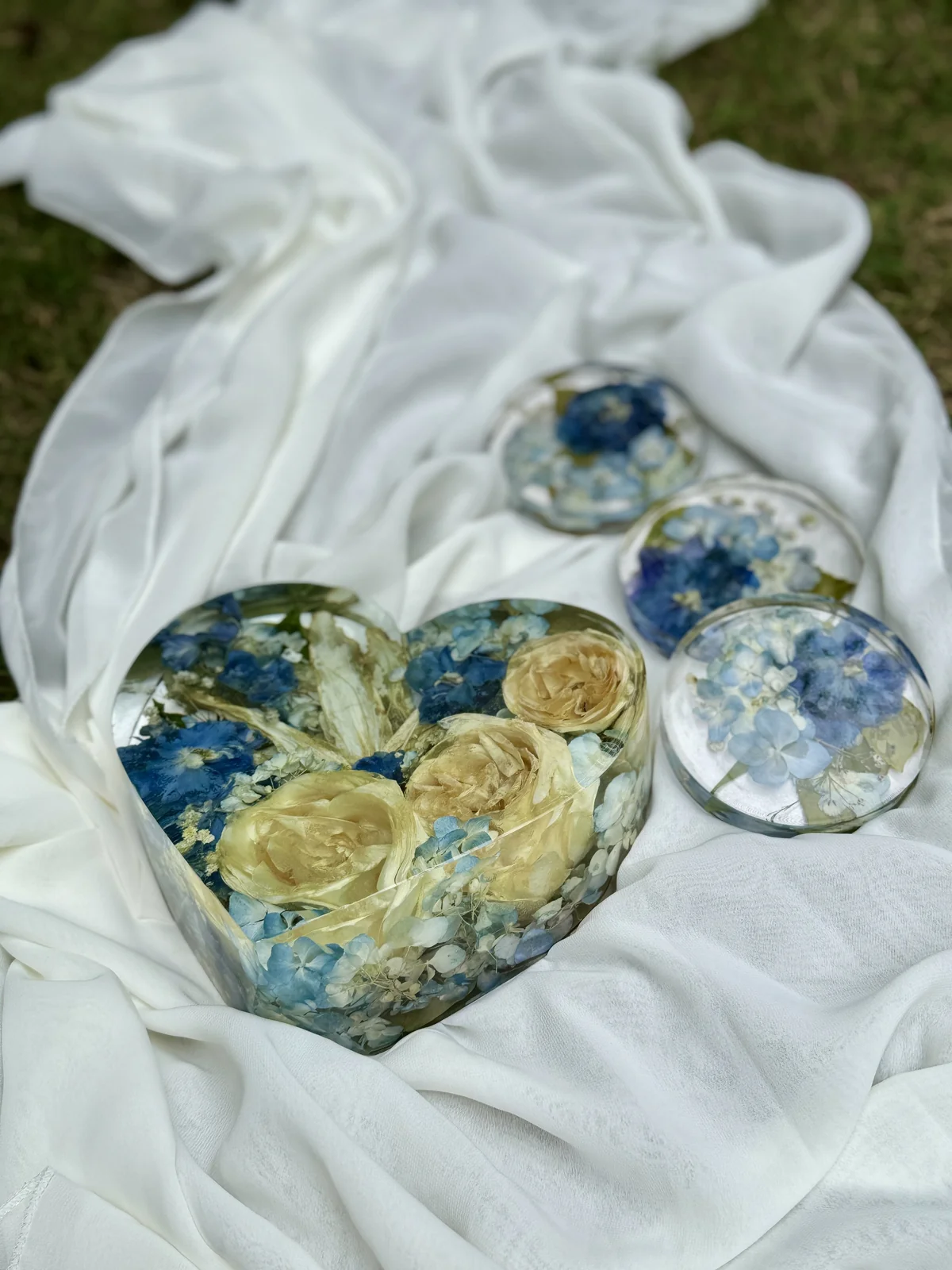 Decorative glass heart and round objects with blue and white flowers resting on white fabric with grass in the background.