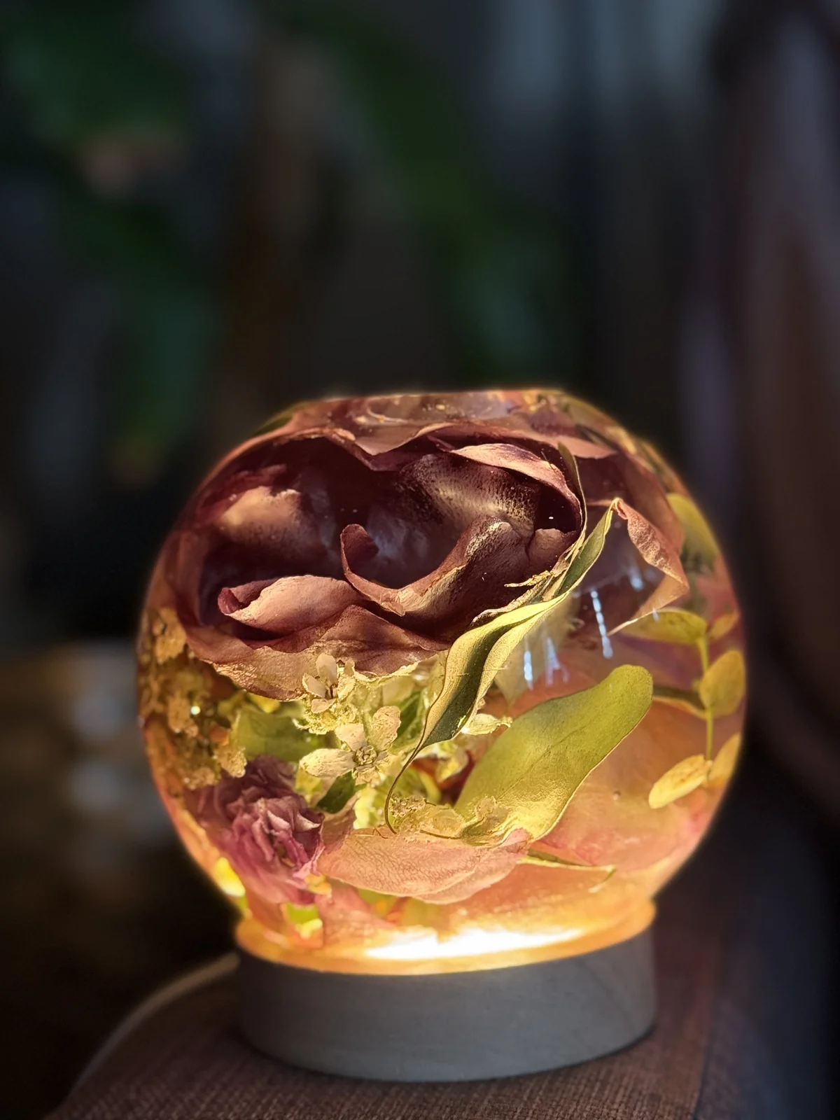 Decorative glass orb lamp with pressed flowers inside, glowing softly on a dark surface.