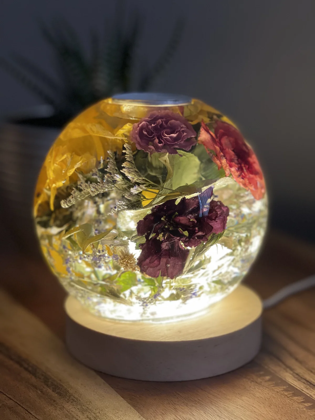 A round resin lamp/candle holder with colourful dried flowers, sitting on a wooden surface with a light base underneath, with a dark background.