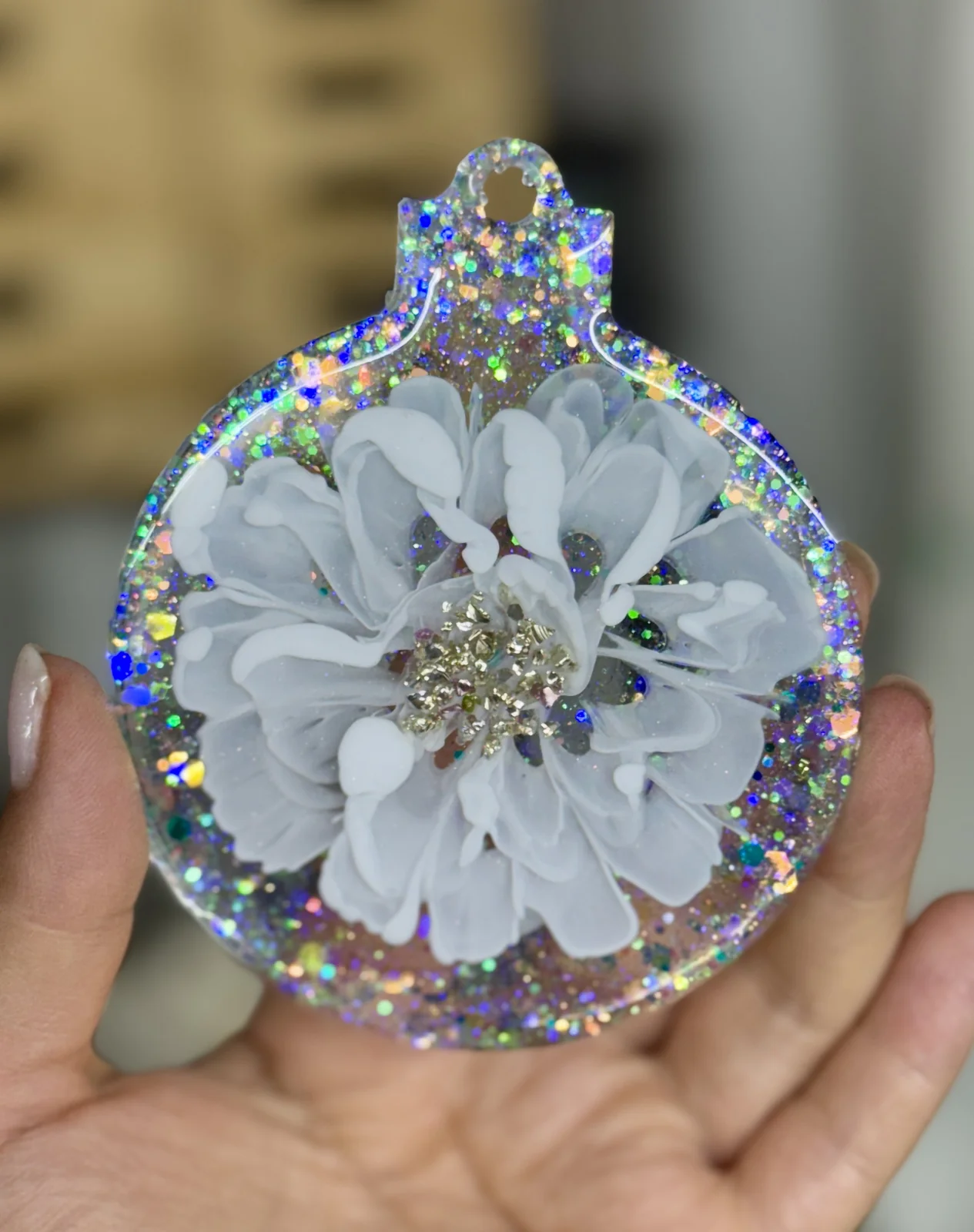 A white floral ornament with iridescent rhinestones and glitter on a transparent background, held in a person's hand.
