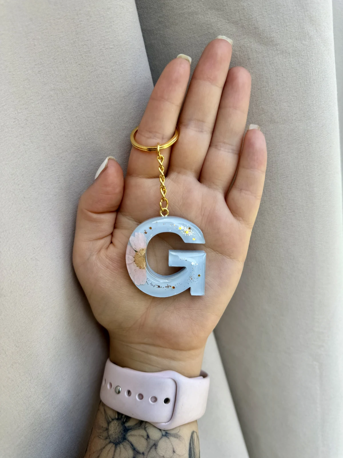 A hand holding a keychain with a large, blue, resin letter 'G' decorated with dried flowers and gold specks. The hand has a gold ring on the middle finger and a light pink watch on the wrist, with a floral tattoo visible on the forearm.
