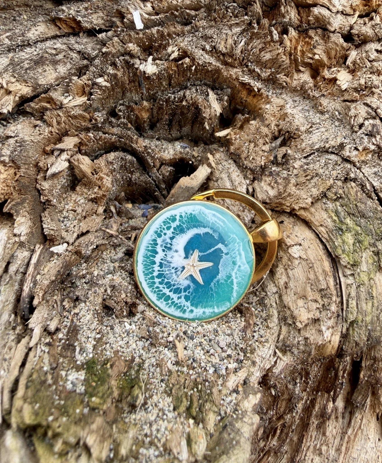 A gold ring with a circular top featuring a beach scene with a starfish, waves, and sand, placed on a textured piece of tree bark.