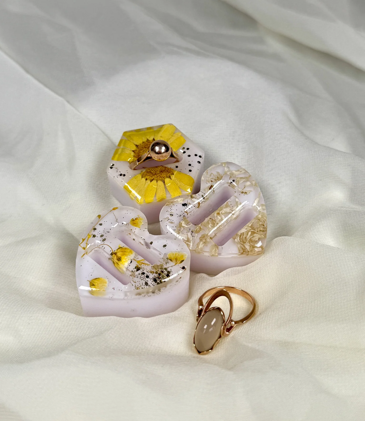 Decorative soap bars with floral and gold accents, along with jewelry pieces including a ring with a pearl and a ring with a beige stone, arranged on a white fabric background.