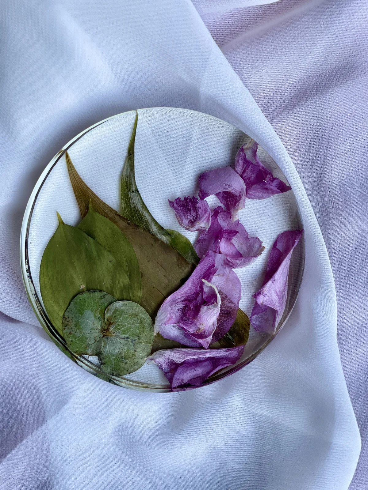 A clear glass bowl filled with green leaves and purple flower petals, placed on a white cloth background.